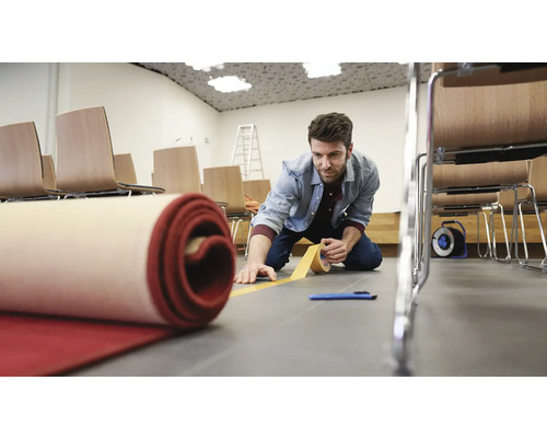 Ein Mann verlegt einen roten Teppich mit Klebeband in einem Raum mit Stühlen und einer Leiter.