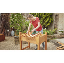 Frau schleift Holz auf einem Hochbeet mit einem Schleifgerät im Garten.