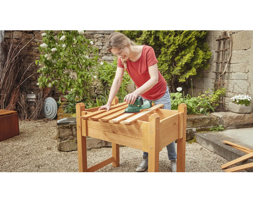 Frau schleift Holz auf einem Hochbeet mit einem Schleifgerät im Garten.