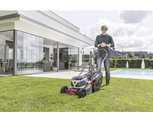 Ein Mann benutzt einen Akku-Rasenmäher in einem Garten mit modernem Haus und Pool.