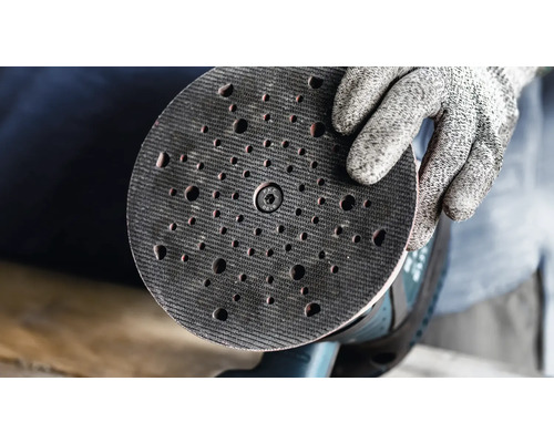 Ein Schleifteller für einen Exzenterschleifer wird von einer Hand mit Schutzhandschuh gehalten.