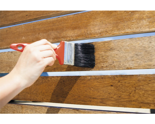 Eine Hand trägt eine Bürste auf einem Holzdeck auf.