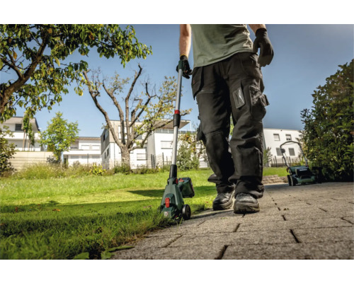 Ein Mann benutzt einen Akku-Rasentrimmer im Garten, ein Rasenmäher steht im Hintergrund.