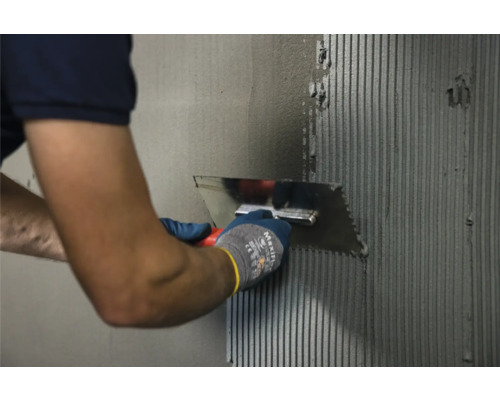 Eine Person trägt mit einer Spachtel Klebstoff auf eine Wand auf.