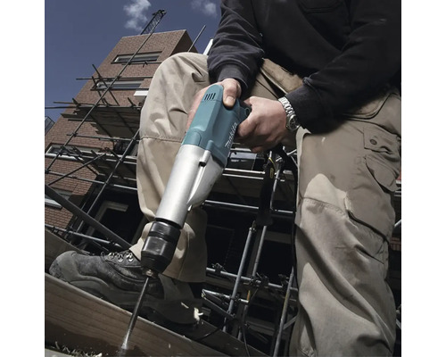 Ein Handwerker bohrt mit einer Bohrmaschine ein Loch in ein Holzbrett auf einem Baugerüst vor einem Gebäude.