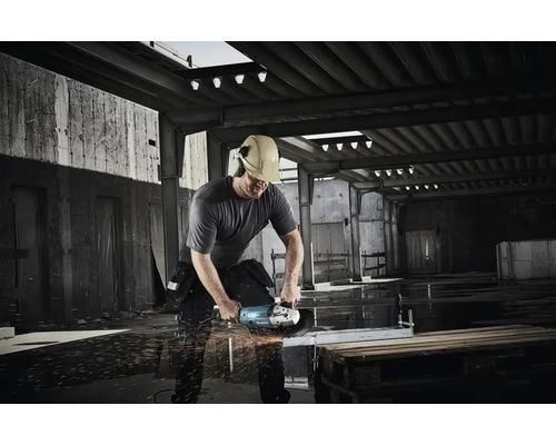 Ein Mann mit Schutzhelm und Gehörschutz schneidet mit einem Winkelschleifer ein Metallstück in einer Werkshalle.