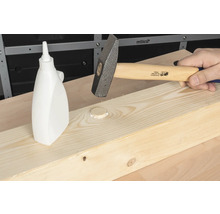 Ein Hammer schlägt einen Holzdübel in ein Holzbrett, daneben eine Flasche mit Holzleim.