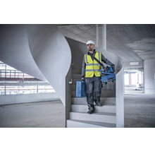 Ein Bauarbeiter mit Schutzhelm und Sicherheitsweste trägt eine Werkzeugkiste und einen Staubsauger die Treppe hinunter.