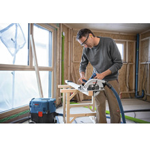 Ein Mann sägt Holz mit einer Handkreissäge in einer Baustelle. Im Hintergrund sind ein Fenster und ein Staubsauger zu sehen.