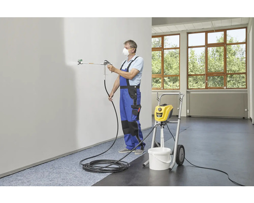 Ein Mann mit Atemschutzmaske sprüht mit einem Farbsprühgerät Farbe an eine Wand. Im Hintergrund steht ein Farbspritzgerät auf einem Rollwagen.