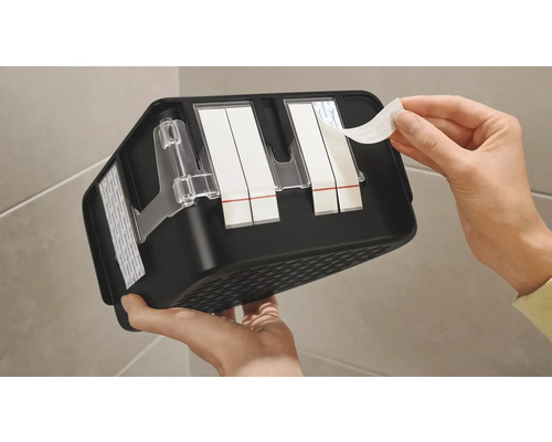 Person entfernt Schutzfolie von Klebestreifen an einer schwarzen Ablage für Badezimmeraccessoires.