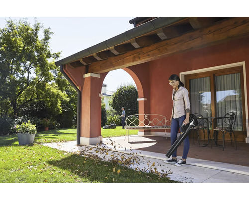 Frau benutzt Laubbläser auf Terrasse im Garten, ein Mann arbeitet mit einem Rasenmäher