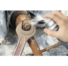 Eine Person sprüht Rostlöser auf eine Mutter, die mit einem Schraubenschlüssel gehalten wird.