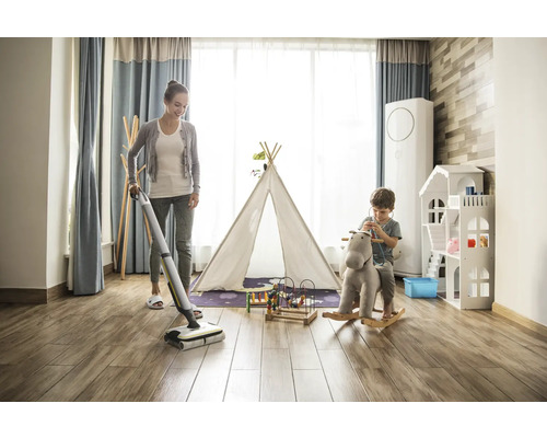 Eine Frau wischt den Holzboden in einem hellen Kinderzimmer mit einem Akku-Wischer, während ein Kind auf einem Schaukelpferd spielt.