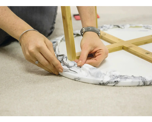Eine Person schneidet überschüssiges Verpackungsmaterial von einem Tisch mit Marmoroptik und Holzbeinen ab.