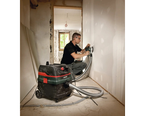 Handwerker schleift Wand mit einem Langhalsschleifer und einem Metabo Staubsauger in einem Raum.