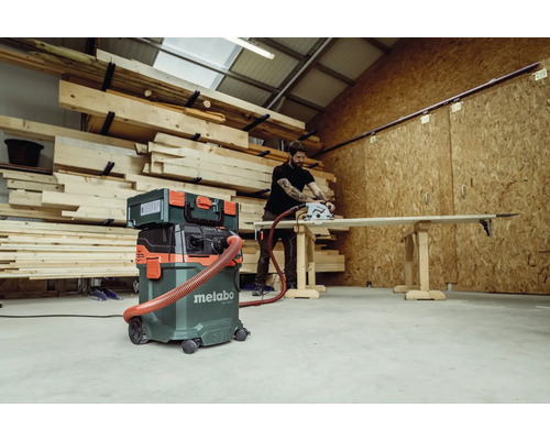 Werkstatt Szene mit Holzlager, Metabo Industriesauger, Werkzeugkoffer und Mann beim Holzsägen