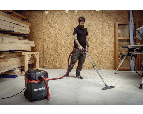 Werkstatt mit Mann, der einen Metabo Staubsauger benutzt, um den Boden zu reinigen, mit Holzstapeln im Hintergrund.