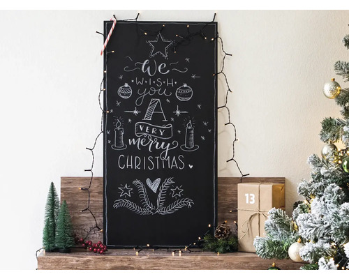 Dekorative Tafel mit Kreidezeichnung und Weihnachtsbeleuchtung auf einem Holzregal, daneben ein Weihnachtsbaum mit Schmuck.