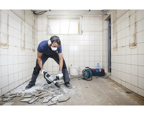 Mann mit Presslufthammer bei Renovierungsarbeiten in einem Raum mit Fliesen an Wänden und Boden
