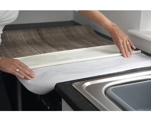 Person bringt selbstklebende Dekorfolie mit Holzoptik auf einer Arbeitsplatte an.