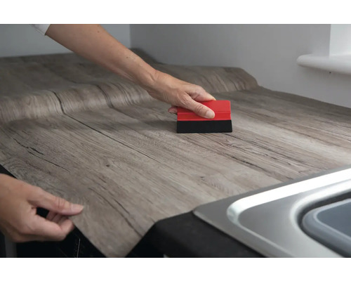 Eine Person bringt eine selbstklebende Holzdekorfolie auf einer Arbeitsplatte an und verwendet einen Rakel.