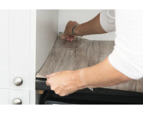 Eine Person bringt eine selbstklebende Folie mit Holzoptik in einem Küchenschrank an.