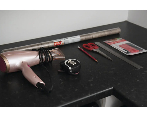 Haartrockner, Maßband, Schere, Bleistift, Lineal und Möbelfolie auf einer Arbeitsfläche.