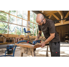 Ein Mann bearbeitet ein Holzwerkstück mit einem Multifunktionswerkzeug und Staubsauger in einer Werkstatt.