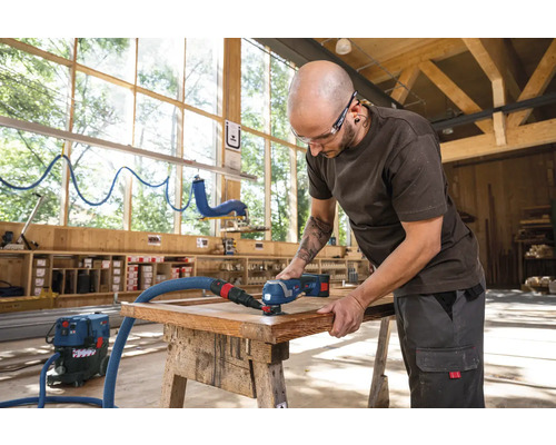 Ein Mann bearbeitet ein Holzwerkstück mit einem Multifunktionswerkzeug und Staubsauger in einer Werkstatt.