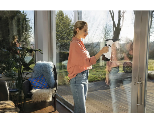 Frau reinigt Fenster mit einem Fensterreiniger in einem hellen Raum.