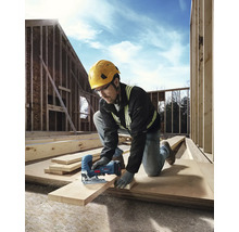 Ein Handwerker mit Schutzhelm und Sicherheitsbrille sägt ein Holzbrett mit einer Stichsäge auf einer Baustelle.