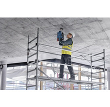 Ein Arbeiter steht auf einem Gerüst und bohrt mit einer Bohrmaschine in eine Betondecke.