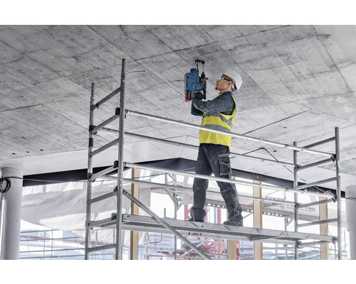 Ein Arbeiter steht auf einem Gerüst und bohrt mit einer Bohrmaschine in eine Betondecke.
