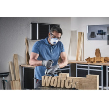 Mann arbeitet mit einer Holzfräse an einem Holzstück mit dem Schriftzug Wolfer, trägt dabei eine Schutzmaske und Schutzbrille in einer Werkstatt.