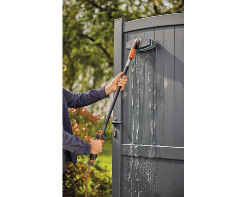 Person reinigt ein graues Gartentor mit einer Waschbürste und Wasserschlauch.