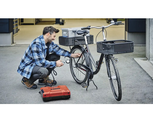 Mann pumpt Fahrradreifen mit einem Kompressor auf. Das Fahrrad hat Körbe und einen Gepäckträger.