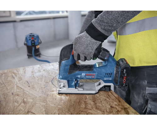 Eine Person sägt mit einer Stichsäge ein Loch in ein Holzbrett. Im Hintergrund ist ein Staubsauger zu sehen.