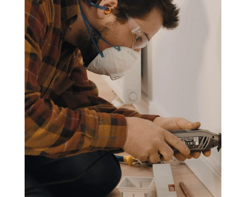 Ein Mann arbeitet mit einem Multifunktionswerkzeug an einer Fußleiste, geschützt mit Atemschutzmaske und Schutzbrille.