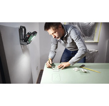 Ein Mann zeichnet mit Bleistift Umrisse auf eine grüne Oberfläche, beleuchtet von einer tragbaren Arbeitsleuchte an der Wand.