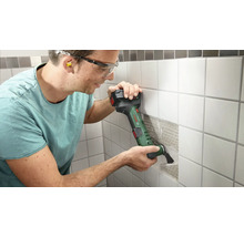 Mann arbeitet mit einem Multifunktionswerkzeug an einer gefliesten Wand. Er trägt dabei eine Schutzbrille und Ohrstöpsel.