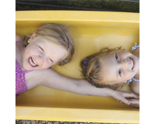 Zwei lachende Mädchen liegen in einem gelben Sandkasten.