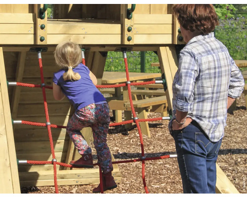 Kleines Mädchen klettert auf ein Spielgerät mit rotem Seilnetz, während eine ältere Frau zusieht.