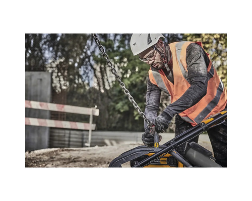 Ein Bauarbeiter mit Helm und Schutzweste bedient eine Rüttelplatte mit einer Kette an einer Baustelle.