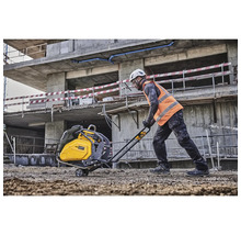 Ein Bauarbeiter bedient eine Rüttelplatte auf einer Baustelle vor einem unfertigen Betongebäude.