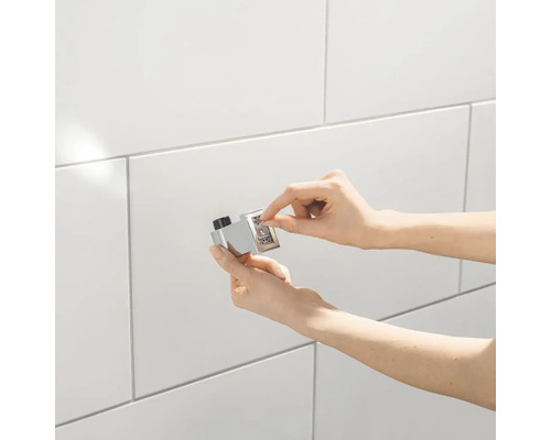 Nahaufnahme einer Hand, die einen verchromten Thermostat-Druckknopf mit Zahlen auf einer gefliesten Wand bedient