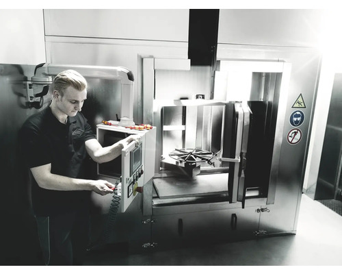 Ein Mann bedient eine Industriewaschmaschine in einer Werkstatt. Sicherheitszeichen weisen auf mögliche Gefahren hin.