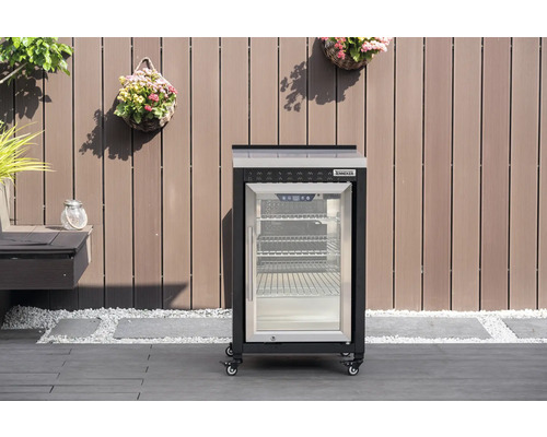 Mobiler Tenneker Outdoor-Kühlschrank mit Glastür und Rollen auf einer Terrasse vor einer Holzwand.