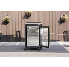 Schwarzer Tenneker Outdoor-Kühlschrank mit geöffneter Glastür und Gitterböden auf einer Terrasse vor einem Sichtschutzzaun.