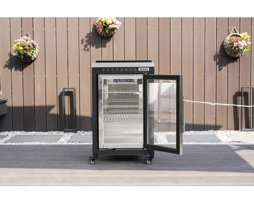 Schwarzer Tenneker Outdoor-Kühlschrank mit geöffneter Glastür und Gitterböden auf einer Terrasse vor einem Sichtschutzzaun.